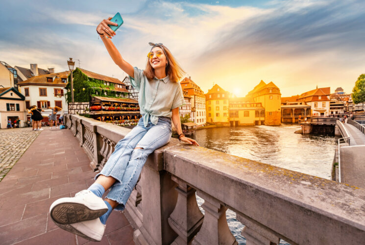 Happy and cheerful Asian girl traveler takes a selfie on the Saint Martin bridge in the Petit France area in Strasbourg. Colorful and dramatic sunset at the background